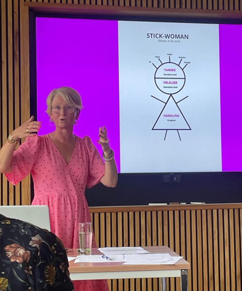 lyshåret kvinde i lyserød kjole, der holder foredrag foran en helt lyserød skærm