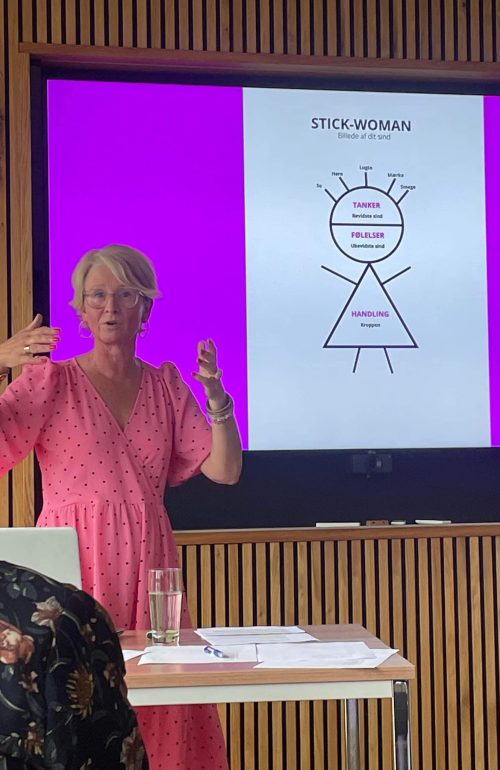 lyshåret kvinde i lyserød kjole, der holder foredrag foran en helt lyserød skærm